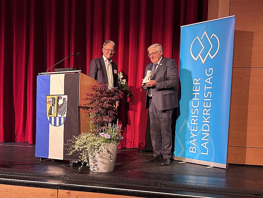 Zwei Männer auf einer Bühne vor rotem Vorhang – einer hält eine Auszeichnung, der andere steht am Rednerpult mit Wappenflagge; rechts Banner mit „BAYERISCHER LANDKREISTAG“, davor zwei Pflanzkübel