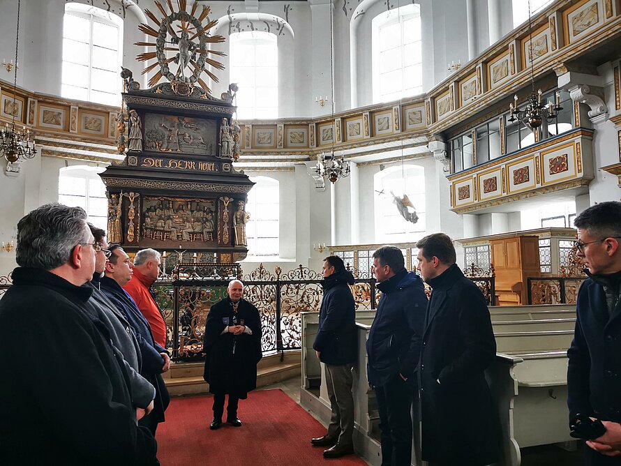 Eine Gruppe von Personen steht in einer reich verzierten Kirche vor einem Altar mit der Inschrift „DIE LIEBE HÖRET NIMMER AUF“; die Szene wirkt wie eine Führung oder ein gemeinsamer Besuch in einem historischen Sakralraum.