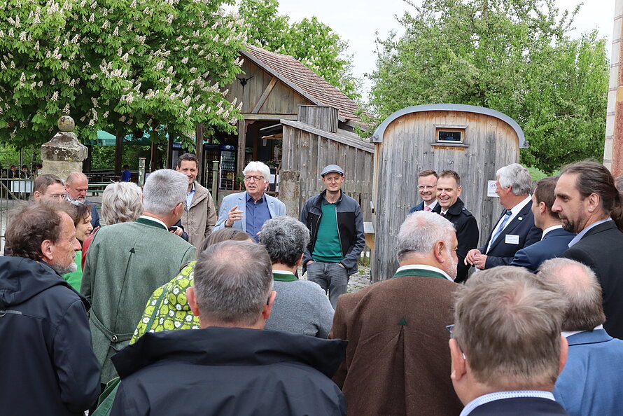Gruppe älterer Personen steht im Freien in einem halbkreisförmigen Arrangement – ein Mann mit weißem Haar spricht, umgeben von Holzgebäuden mit Ziegeldach und grüner Vegetation; Kleidung teils formell, Umgebung ländlich geprägt.