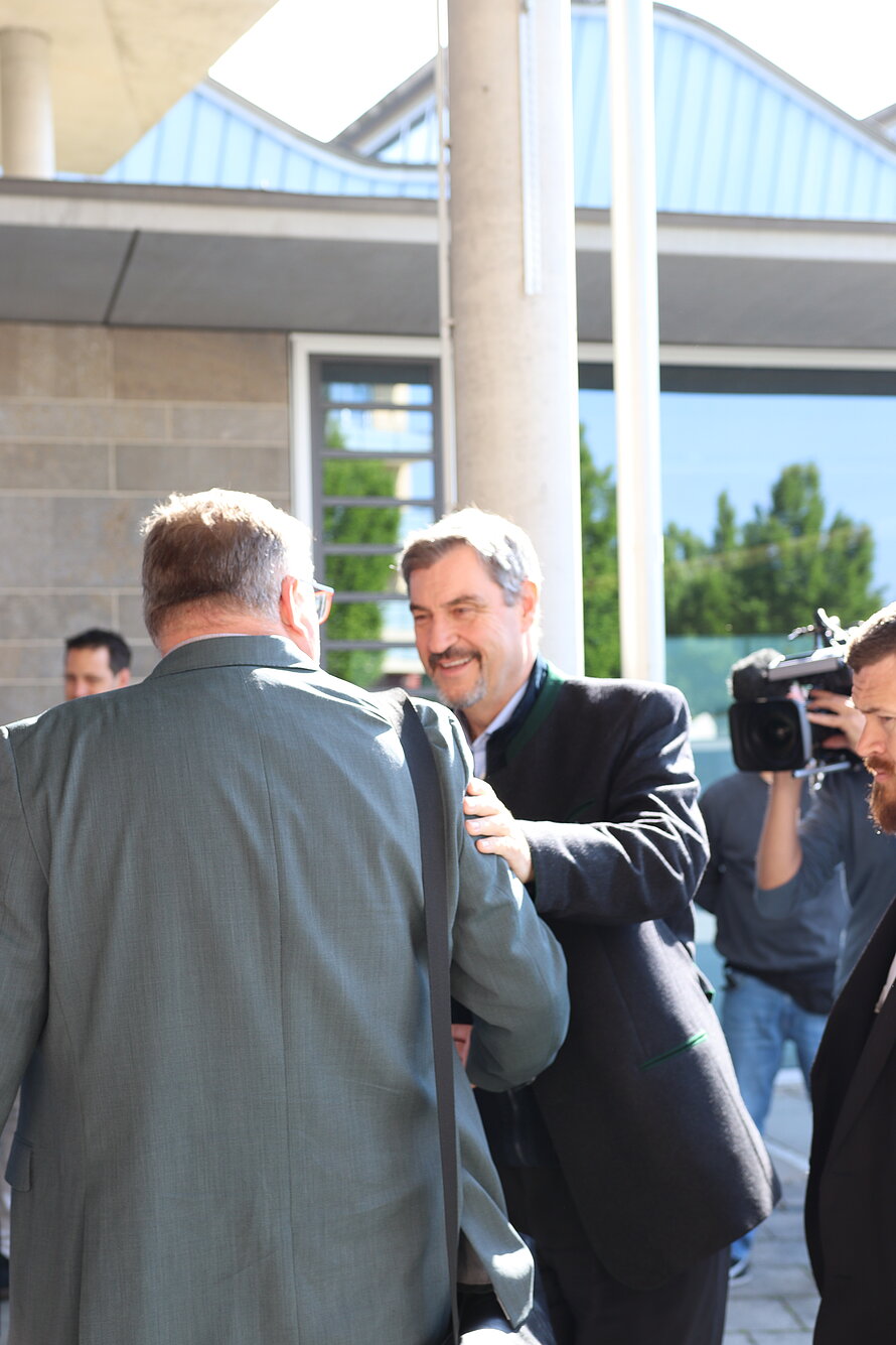 Zwei Personen im Anzug begrüßen sich per Handschlag im Außenbereich vor einem Gebäude mit großen Fenstern und Betonsäule – im Hintergrund dokumentiert ein Kameramann die Szene mit professionellem Equipment; weitere Personen sind anwesend.
