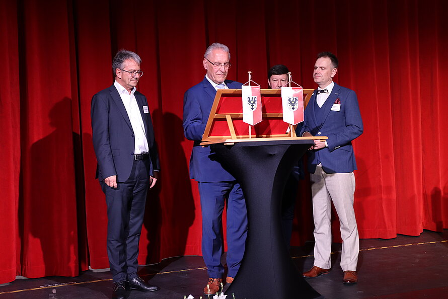 Vier Männer in Anzügen stehen auf einer Bühne mit rotem Vorhang – sie versammeln sich um einen schwarzen Stehtisch mit Holzrahmen, darin zwei kleine Flaggen mit schwarzem Doppeladler auf weißem Grund; zentrale Person führt eine formelle Handlung aus, möglicherweise Präsentation oder Unterzeichnung.