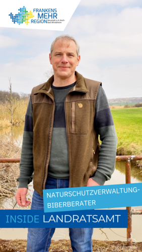 Mann in Outdoorkleidung vor rostigem Geländer in ländlicher Umgebung – Wiese, Wasserlauf und teils kahle Bäume im Hintergrund