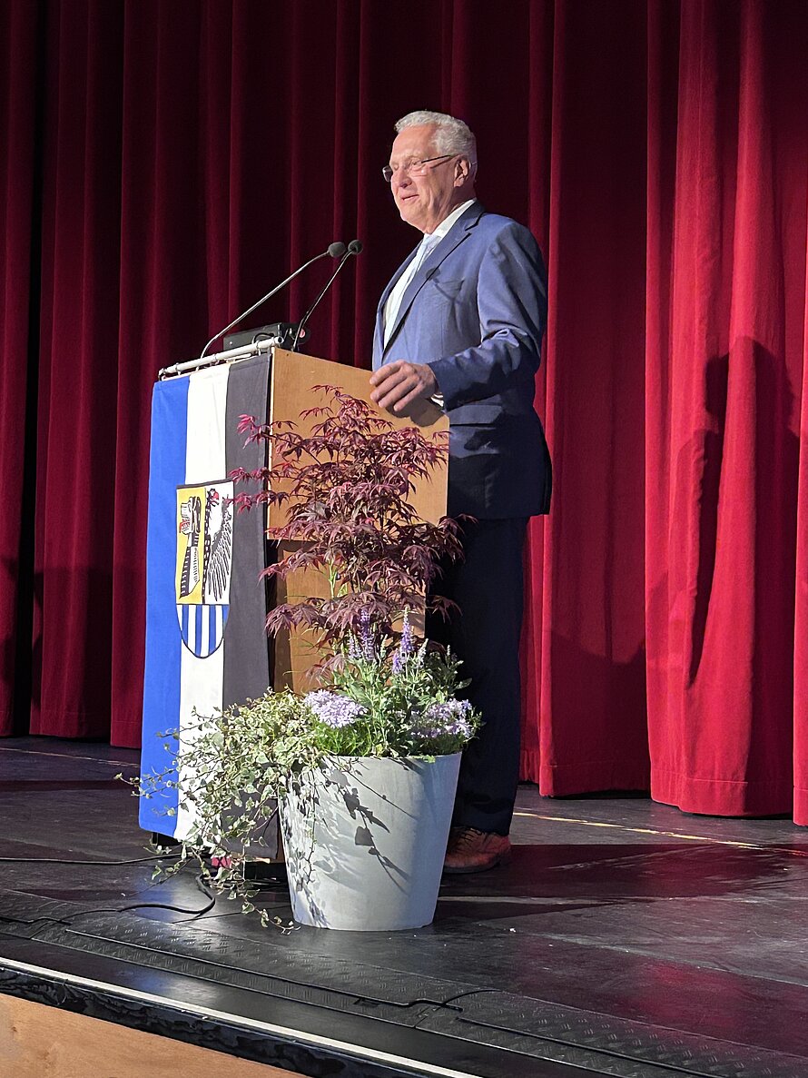 Älterer Mann im blauen Anzug steht hinter einem mit Blumen geschmückten Rednerpult auf einer Bühne mit rotem Vorhang – Mikrofon montiert, Pult mit Banner samt stilisiertem Adler und Streifenmotiv versehen; Szene zeigt eine formelle Ansprache in institutionellem Rahmen.