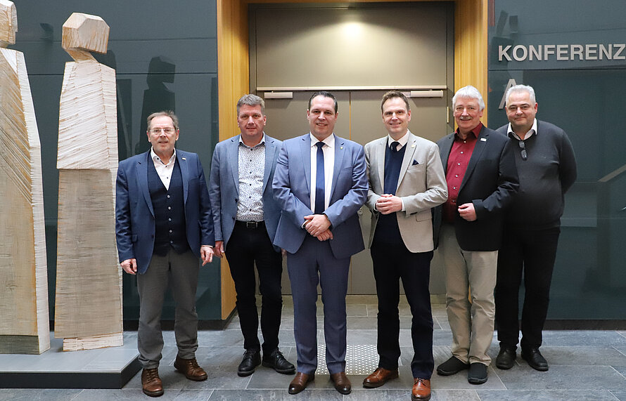 Sechs Personen stehen nebeneinander in einem Konferenzraum vor einer Wand mit dem Schriftzug „KONFERENZ“ und zwei abstrakten Holzskulpturen; sie tragen geschäftliche Kleidung und posieren für ein Gruppenfoto.