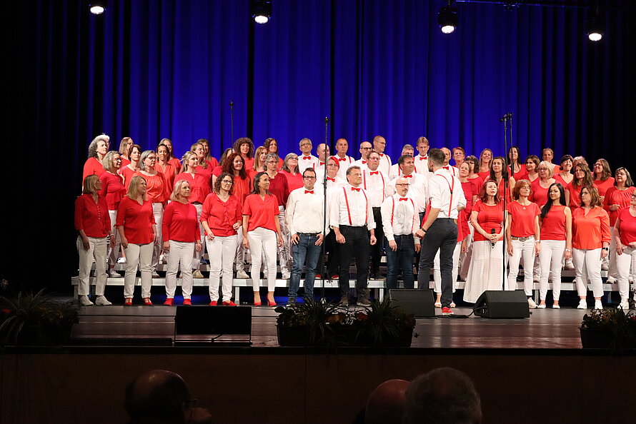 Großer gemischter Chor auf beleuchteter Bühne mit blauer Rückwand – Frauen in roten Oberteilen und weißen Hosen oder Röcken, Männer in weißen Hemden mit roten Hosenträgern und Fliegen; gestaffelte Anordnung auf Podesten, deutet auf eine organisierte musikalische Aufführung.