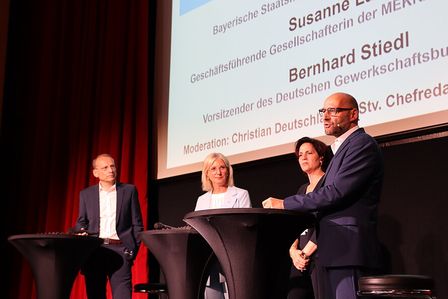 Vier Personen in Businesskleidung stehen auf einer Bühne hinter hohen Stehtischen – im Hintergrund Leinwand mit teils abgeschnittenem Text zu einer Veranstaltung der Bayerischen Staatsregierung; genannte Namen: Susanne D. (MERK), Bernhard Stiedl (Deutscher Gewerkschaftsbund), Moderation durch Christian Deutschl. Bühne und Setting deuten auf eine formelle Diskussionsrunde.