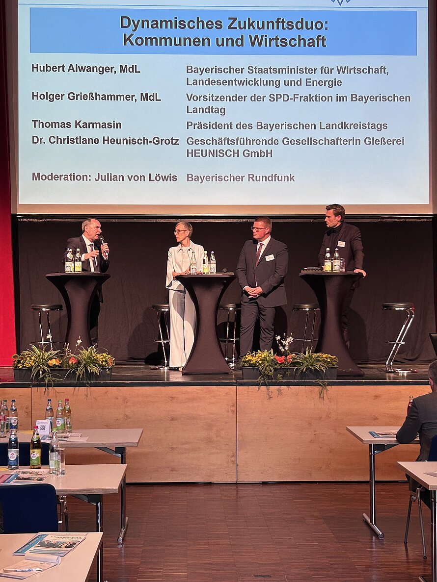 Podiumsdiskussion mit fünf Personen an einem Tisch mit Mikrofonen – hinter ihnen eine Leinwand mit Titel „Dynamisches Zukunftsduo: Kommunen und Wirtschaft“ sowie Namen und Funktionen der Teilnehmenden: Hubert Aiwanger, Holger Greilhammer, Thomas Karmasin, Georg Huber, Moderation durch Julian von Löwis; Bühne und Setting klar auf eine offizielle Veranstaltung