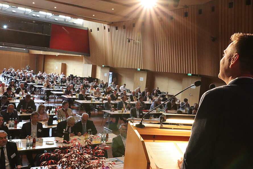 Blick von hinten auf eine Person am Rednerpult in einem großen Saal – Publikum sitzt an Tischen mit Wasserflaschen und Unterlagen, Mikrofone und Technik am Pult sichtbar; Raum mit Holzverkleidung und hoher Decke, helle Lichtquelle von oben beleuchtet Bühne und Teile des Auditoriums.