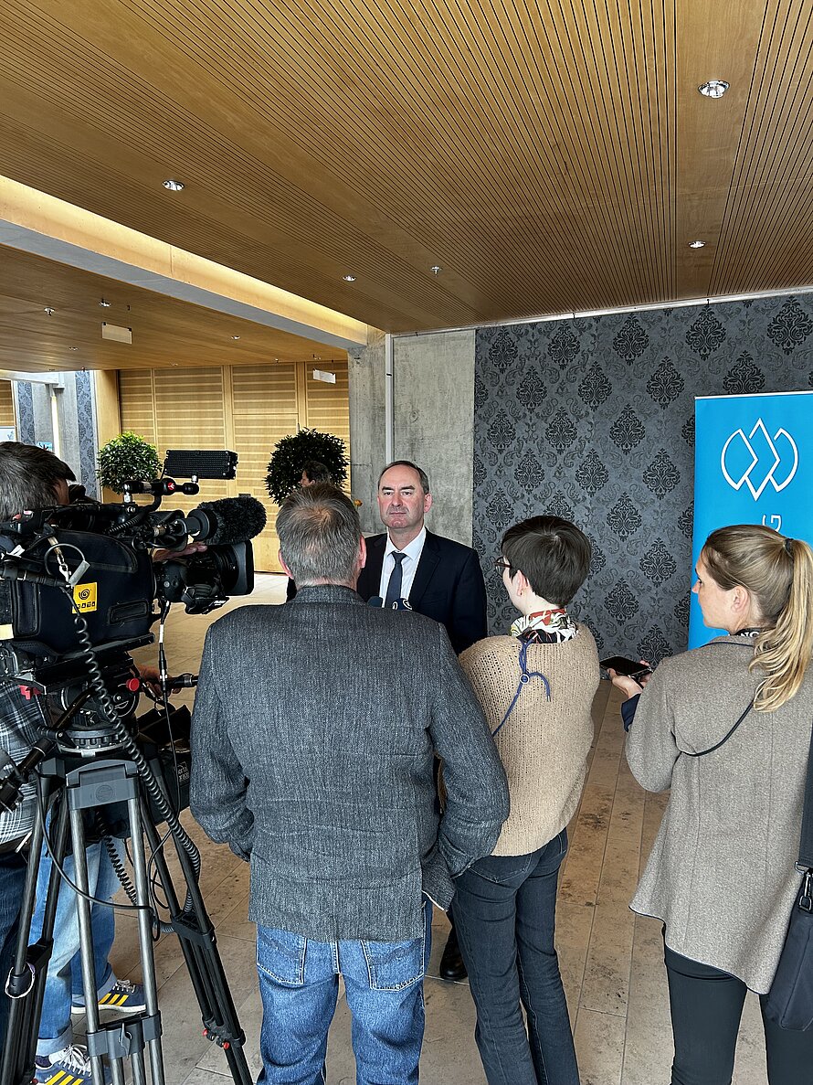 Mann im Anzug spricht vor mehreren Personen mit Mikrofonen und Kameras – professionelle Aufnahmegeräte und Innenraum mit Holzdecke und gemusterter Wand im Hintergrund; blaues Schild mit weißem Logo sichtbar. Szene zeigt eine medienbegleitete Stellungnahme oder Interview.