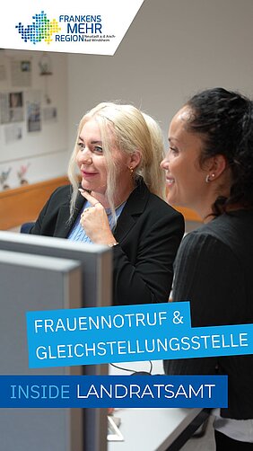 Zwei Personen stehen in einem Büro und betrachten gemeinsam einen Bildschirm – eine trägt einen schwarzen Blazer, die andere hat dunkle Locken und lächelt