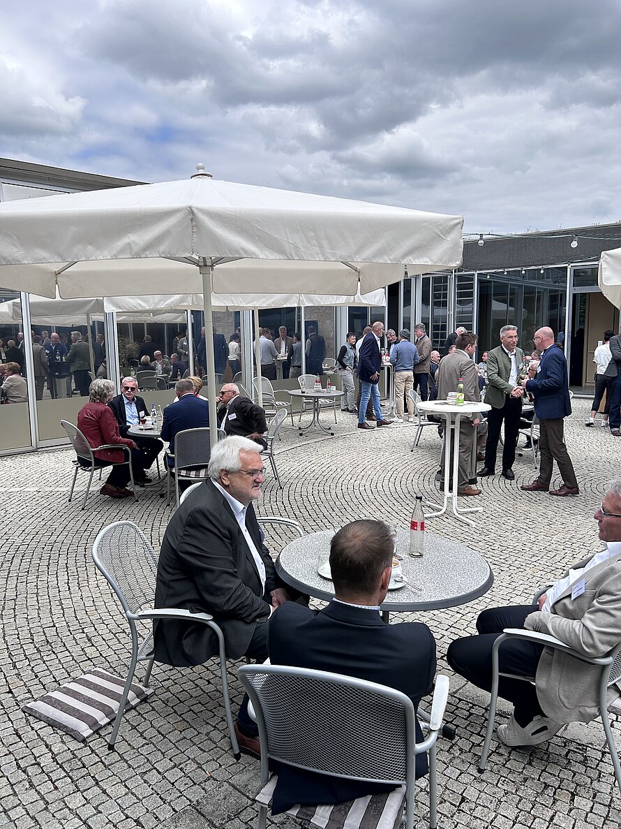 Außenveranstaltung auf gepflasterter Fläche mit weißen Sonnenschirmen – Gäste in formeller Kleidung sitzen und stehen an Metalltischen vor moderner Glasfassade; lockere Gespräche und gedämpftes Licht unter bewölktem Himmel.