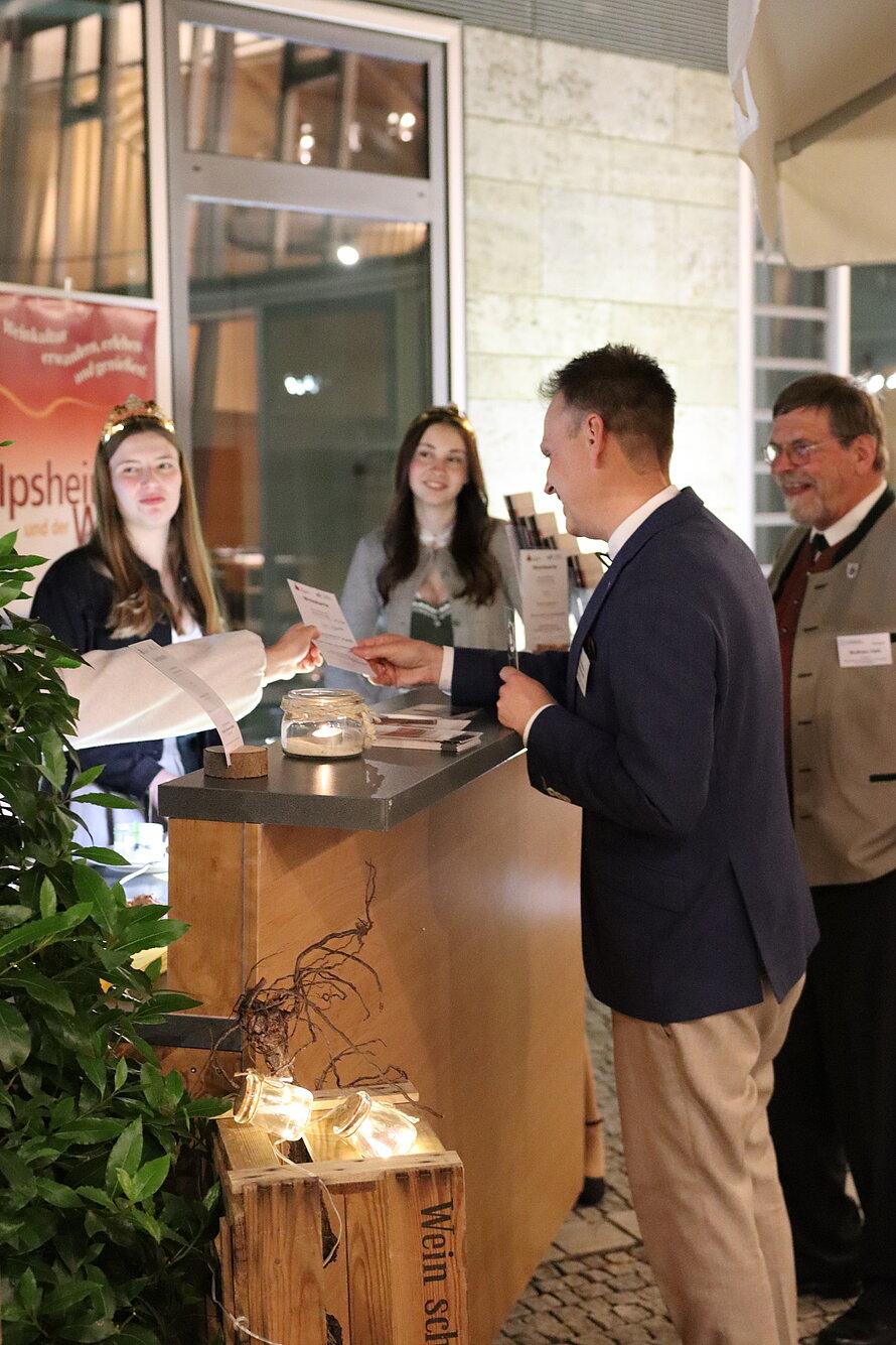 Vier Personen an einem Empfangstisch in einem Innenraum mit großen Fenstern und Fliesenboden – zwei Frauen hinter dem Tisch, eine mit Blumenkranz, übergeben oder empfangen Unterlagen von zwei Männern davor; Tisch dekoriert mit Pflanze und beleuchteter Holzkiste, im Hintergrund Banner mit teilweise lesbarem Schriftzug „Hopsh…“.