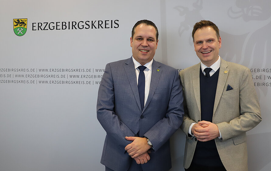 Begrüßung Zwei Personen in Anzügen stehen vor einem Hintergrund mit dem Schriftzug „ERZGEBIRGSKREIS“ und einem Wappen mit Löwe und Bergbausymbol; die Szene wirkt wie ein offizieller Termin oder Empfang.